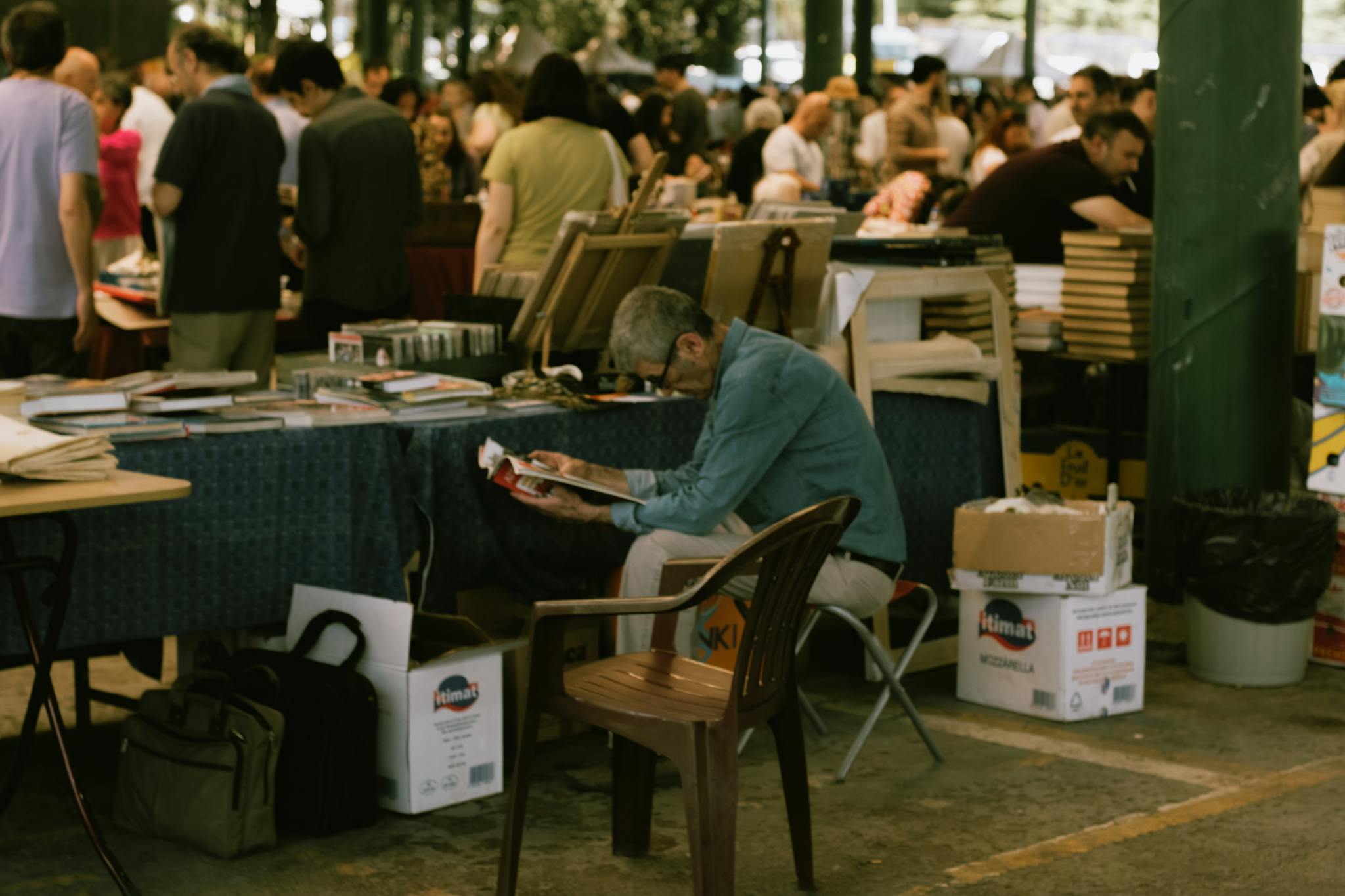 Older man reading a magazine at a bustling outdoor book market.